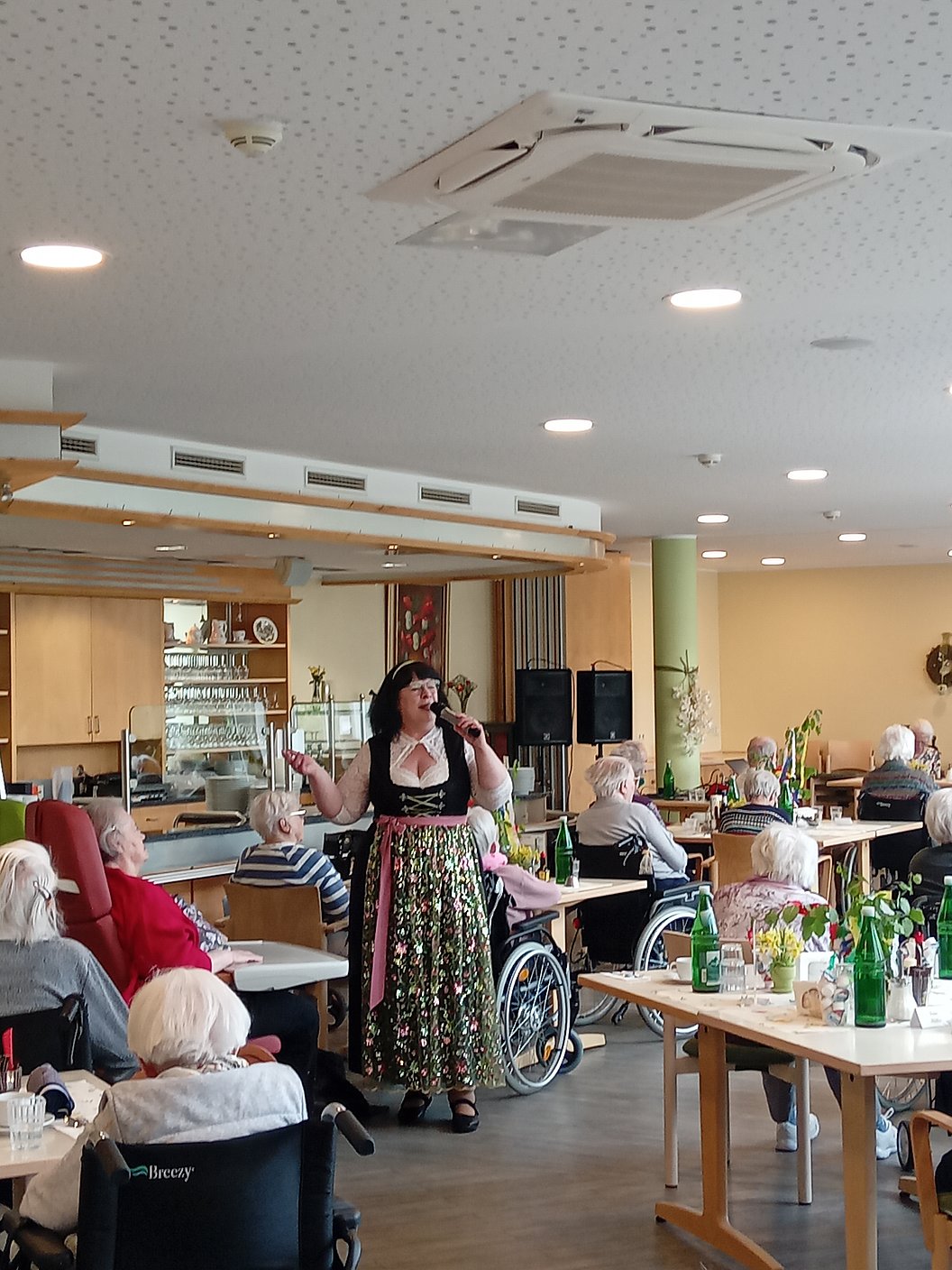Frau in traditioneller Tracht singt mit Mikrofon vor sitzenden älteren Menschen in einem hellen Gemeinschaftsraum mit Tischen und Stühlen