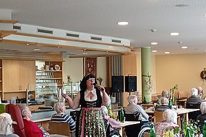 Frau in traditioneller Tracht singt mit Mikrofon vor sitzenden älteren Menschen in einem hellen Gemeinschaftsraum mit Tischen und Stühlen