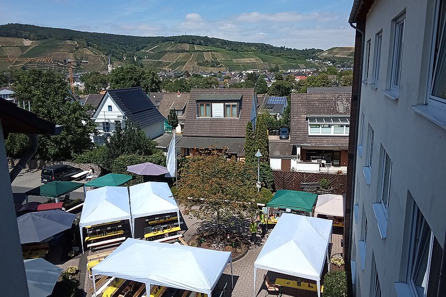 Blick von oben auf einen kleinen Marktplatz mit mehreren weißen und grünen Pavillons vor Wohnhäusern und Weinbergen im Hintergrund