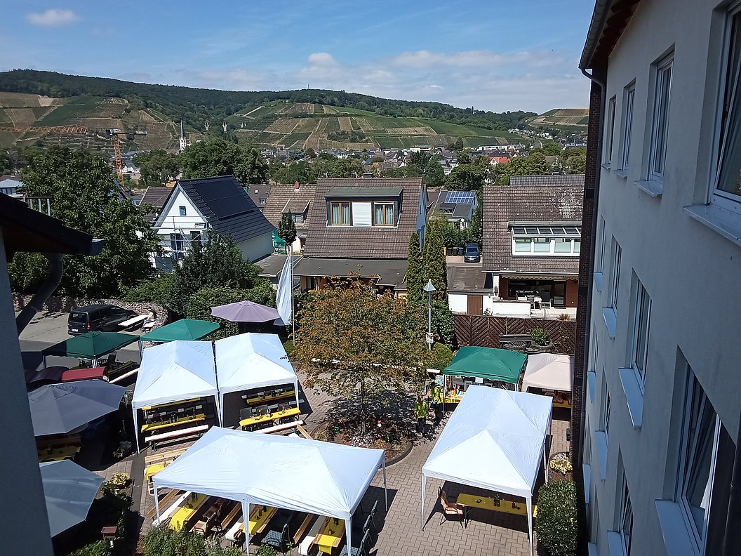 Blick von oben auf einen kleinen Marktplatz mit mehreren weißen und grünen Pavillons vor Wohnhäusern und Weinbergen im Hintergrund