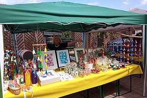 Marktstand mit gelber Tischdecke unter grünem Pavillon, darauf bunte handgefertigte Dekorationsartikel und eine Frau im Hintergrund
